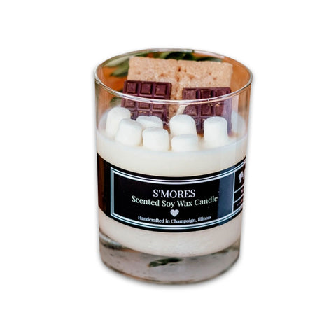 An glass candle holder with a layer of white wax and a piece of chocolate on top, labeled 'S'mores Scented Soy Wax Candle'.