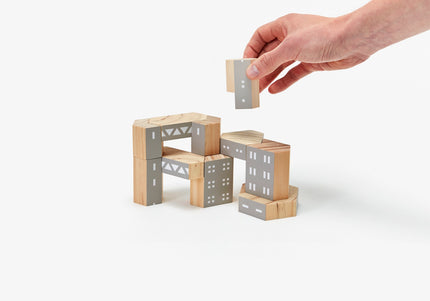 A hand is seen placing a gray block onto a structure made from brown and gray wood building blocks designed to represent architectural brutalism elements. 