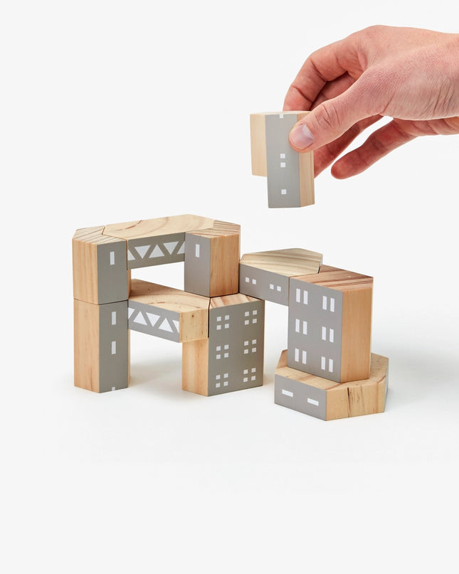 A hand is seen placing a gray block onto a structure made from brown and gray wood building blocks designed to represent architectural brutalism elements. 