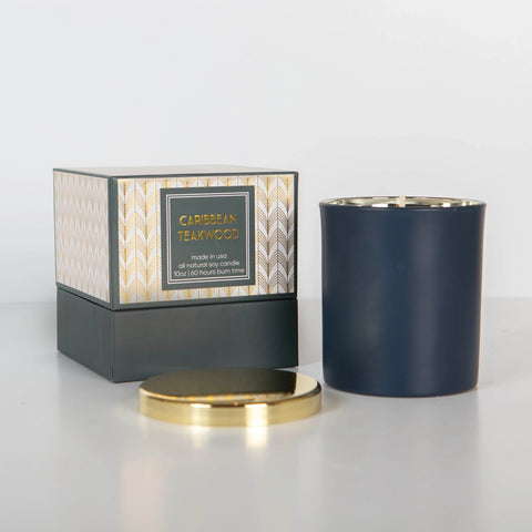 A boxed candle set with a matte blue jar and gold lid alongside its packaging, labeled as 'Caribbean Teakwood'.