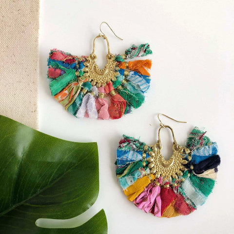 A pair of colorful, fan-shaped earrings made with fringed Kantha textiles, displayed on a white background with a green leaf to the left.