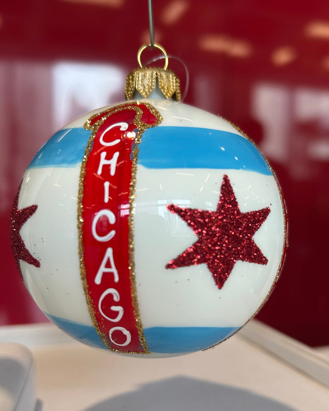 A spherical Christmas ornament featuring the Chicago flag design, with a vertical red banner reading 'CHICAGO' and two red glitter stars, hanging against a red background.