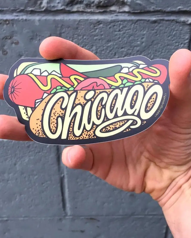 Hand holding a hot dog 'Chicago' sticker against a gray wall.