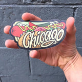 Hand holding a hot dog 'Chicago' sticker against a gray wall.