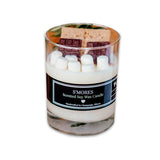 An glass candle holder with a layer of white wax and a piece of chocolate on top, labeled 'S'mores Scented Soy Wax Candle'.