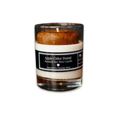A clear glass candle with a brown wax top resembling an apple cider donut, labeled 'Apple Cider Donut Scented Soy Wax Candle'.