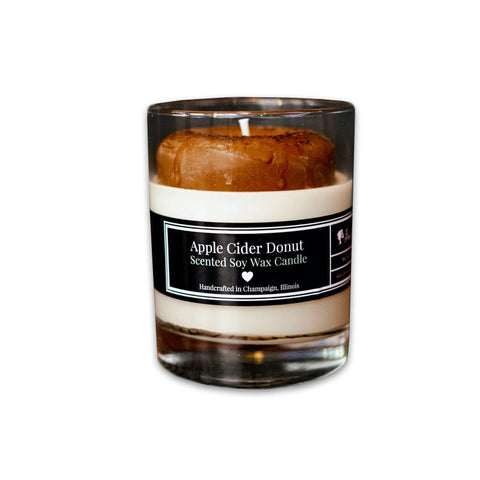 A clear glass candle with a brown wax top resembling an apple cider donut, labeled 'Apple Cider Donut Scented Soy Wax Candle'.